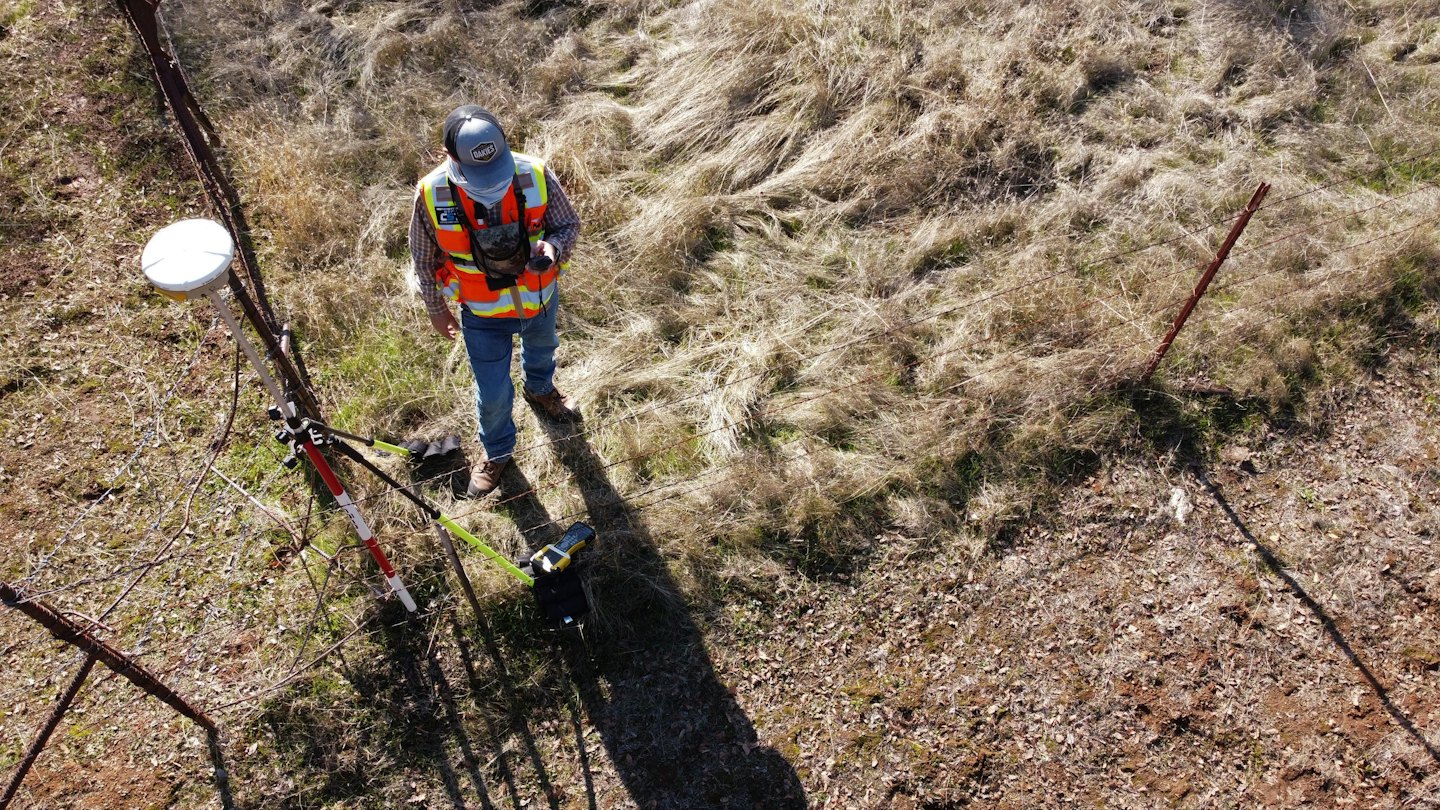 Geodeta obsługuje sprzęt na słonecznym, trawiastym polu.
